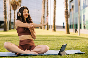 Young pretty woman looking to tablet and doing stretching with headphones. Sitting in lotus pose on blue mat wearing brown sportswear. Yoga time, technology concept 