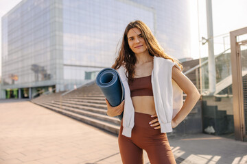 Attractive fit woman staying at street with blue yoga mat in hands. Wearing brown top, leggings and white hoodie , keep hand on side. Concept of workout outdoors 