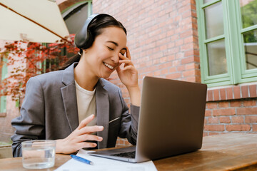 Asian woman in headphones gesturing and working with laptop while sitting in cafe