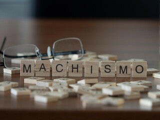 machismo word or concept represented by wooden letter tiles on a wooden table with glasses and a book - obrazy, fototapety, plakaty