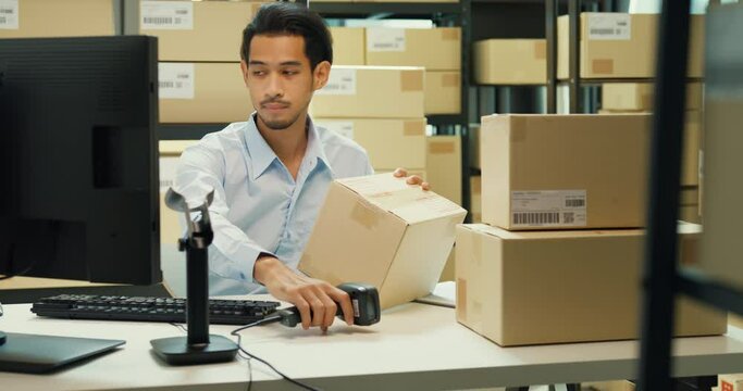 Happy Asian man wear formal uniform shirt sit in front of desk use computer and barcode machine scanner record customer online data in cardboard box delivery at warehouse. Startup business concept.