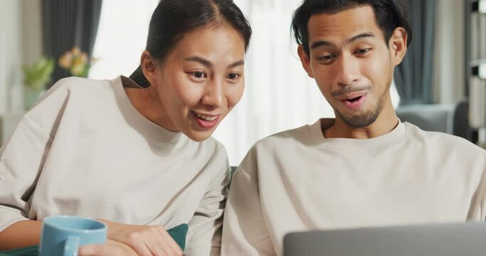 Happy young Asian couple sit on couch use laptop successful payment online shopping at home on holiday. Smile young husband and wife excited at home browsing application on gadgets. Lifestyle concept.