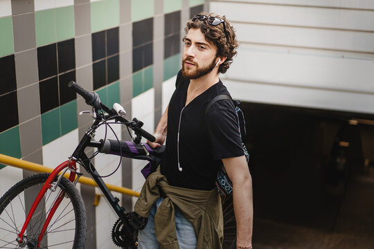 Young adult hipster bearded man with earphones exits the subway station stairs holding his bicycle - people healthy and sustainability lifestyle concept