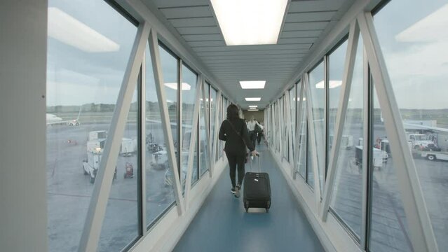 Slow motion footage of a person with a roller bag suitcase walking down the jetway towards a plane.
