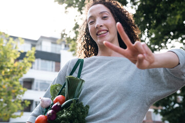 Cheerful woman with bag of vegetables gesturing peace sign