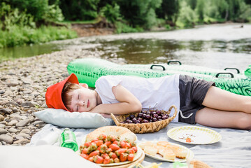 Boy sleeping on picnic blanket by cherry basket at riverbank