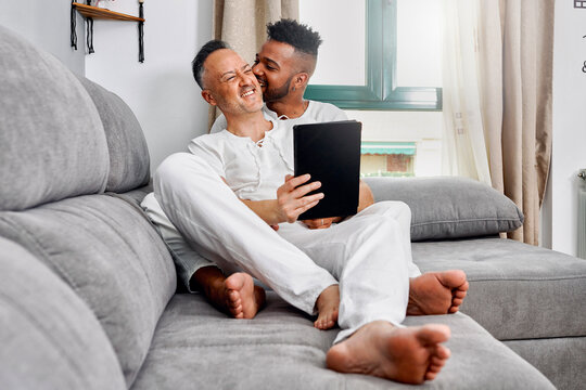Young man biting ear of boyfriend sitting on sofa at home