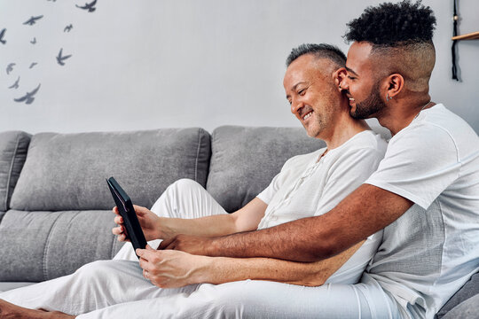 Smiling gay couple sharing tablet PC sitting on sofa at home