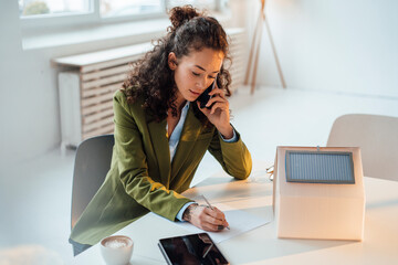 Businesswoman talking on smart phone writing at desk in office