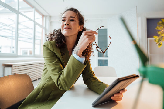 Smiling businesswoman with tablet PC and eyeglasses sitting at desk in office