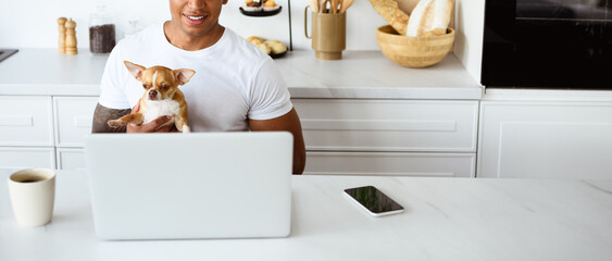 Smiling african american man holding Chihuahua dog near devices and morning coffee in kitchen, banner 