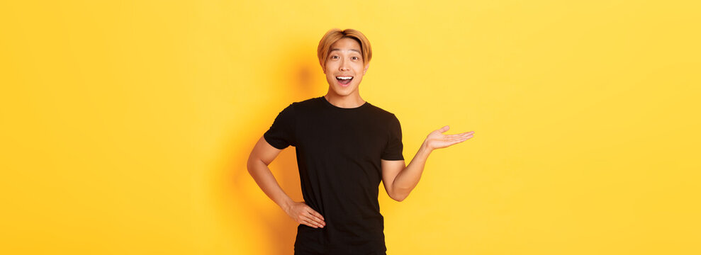 Portrait of happy and excited smiling asian guy holding something on hand over yellow background
