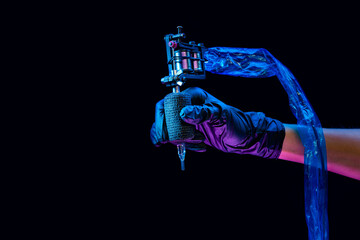 Closeup of tattooer master's hand in black glove holding machine for making tattoo art on body isolated on dark background in neon light. - obrazy, fototapety, plakaty