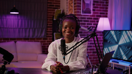 Portrait of african american podcaster sitting at desk in home recording studio with boom arm microphone and digital audio mixer. Online radio host smiling confident at camera while broadcasting live.