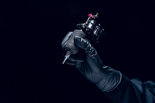 Machine for tattoo. Human hand in black protective glove holding professional equipment for making tattoo on body isolated on dark background.