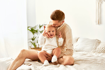    Parental care and love. The father sits on the bed, holds and kisses the laughing baby.