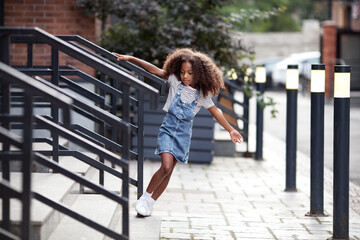 African american kid girl walks down steps on city street. Portrait of child in denim skirt and T-shirt outdoors