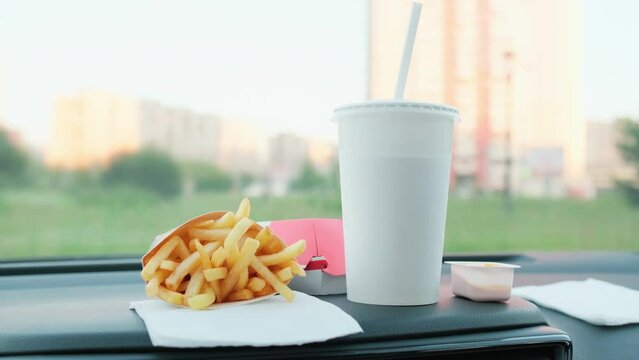 Fast food on a car dash. Burger, french fries and drink with straw in a car. Blurred cityscape on background.