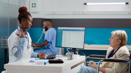 African american doctor doing checkup visit with female wheelchair user, talking about physical disability and healthcare diagnosis. Consulting woman with impairment at medical appointment.