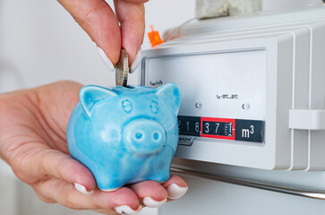 Woman puts coin in piggy bank near a gas meter at home. Natural gas consumption, cost of utilities and saving gas for heating in winter, concept.