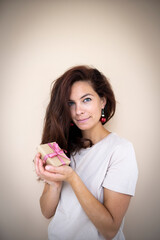 pretty young woman with beautiful long hair is holding a small christmas gift in her hand and has earrings that look like christmas tree baubles standing in front of light brown background