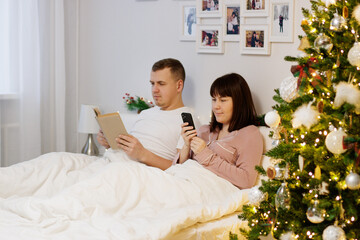 Obraz na płótnie Canvas couple reading book and using smartphone in decorated bedroom with christmas tree