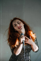 Portrait of redhead female singer woman in sparkly evening dress holding microphone on dark night background. Excited female singer with modern microphone in hands