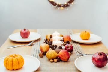 Fall table setting for celebration Thanksgiving day family party. Autumn composition with candles, fruits, nuts and cones. Plates with pumpkins and pomegranate. Natural autumn decor. Selective focus