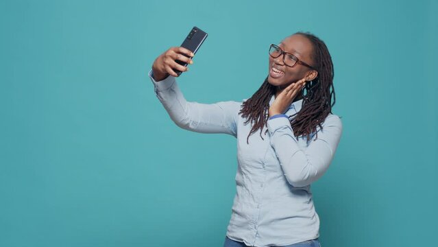 Cheerful happy woman taking pictures on smartphone app, having fun with mobile phone camera to take funny selfies in studio. Natural confident person photographing over blue background.