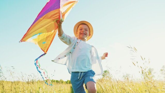 Fast little boy runs across the field with a kite in his hands fluttering in wind over his head. Front view on child walks in nature in summer and has fun play. Family trip out of town to fresh air.
