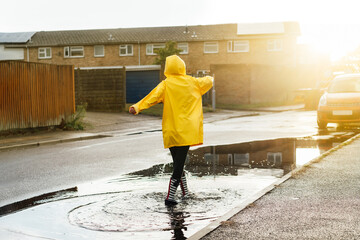 Woman having fun on the street after the rain. Back view woman wearing rain rubber boots and yellow...