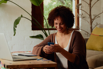 Confident biracial young woman using mobile phone by laptop in living room at home