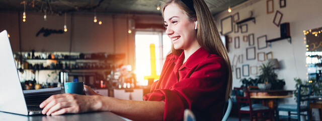 Pretty woman studying with laptop in big bright open space. Wearing red smart-casual dress and drinking coffee