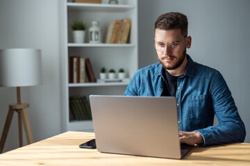 Bearded man in denim shirt working on laptop at home, coding on computer remotely