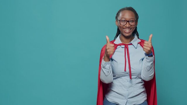 Female superhero with cape doing thumbs up gesture on camera, advertising like and approval sign with red cloak and hero cartoon character costume. Woman giving okay good symbol.