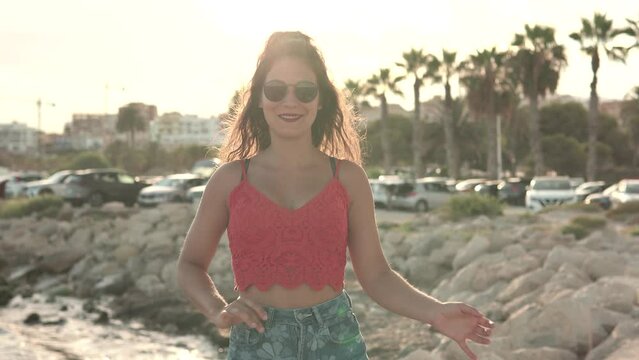 Curly haired brunette woman in red top dancing bachata with the seafront in the background. Video of a young girl dancing latin rhythms.