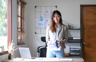 Charming Asian woman with a smile standing holding papers and looking at the camera at the office.