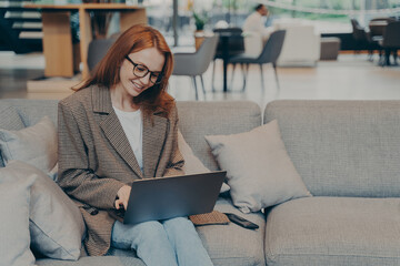 Woman in glasses sitting on cozy couch in office lounge while chatting on computer laptop