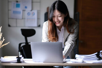 Beautiful Asian businesswoman standing and working on laptop with smiling face while working in office.