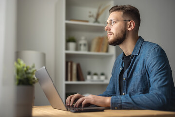 Bearded man in denim shirt working on laptop, coding on computer remotely, bright home interior