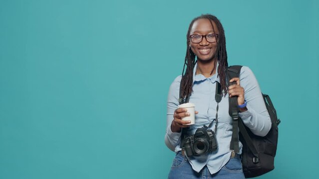African american woman holding coffee cup and backpack before travelling to holiday trip to take photos. Leaving on vacation with camera and photography equipment, taking photos.