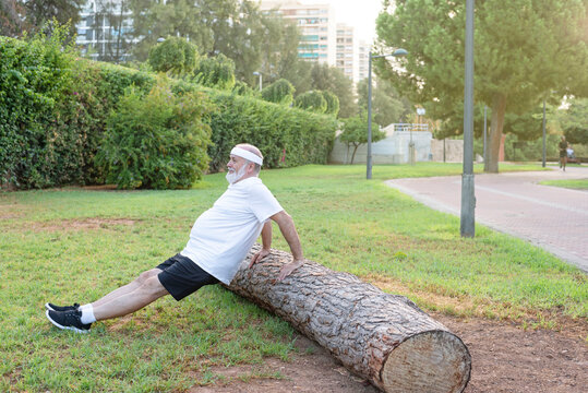 Mature Man Practicing Doing Dips Exercise For Triceps And Biceps Muscle In The Park. Side View Shot Of Elderly Male Exercising With A Log