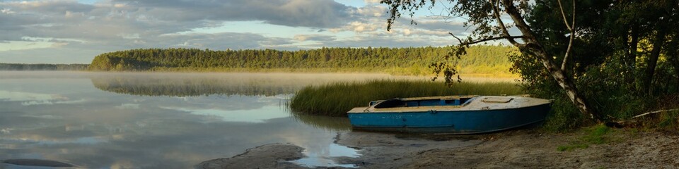 serene morning on a calm forest lake with an old metal boat on the shore, reeds and foggy haze above the water with a cloudy sky reflecting. widescreen panoramic view