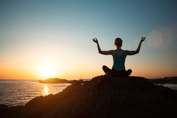 Woman meditating in the lotus position doing yoga by the ocean in amazing sunset.