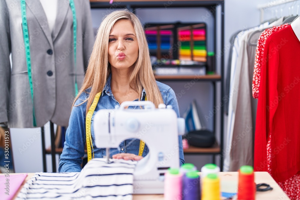 Poster blonde woman dressmaker designer using sew machine looking at the camera blowing a kiss on air being