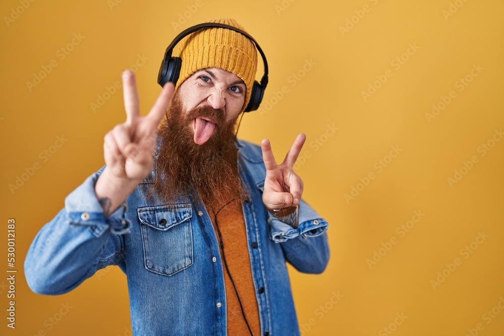 Canvas Prints Caucasian man with long beard listening to music using headphones smiling with tongue out showing fingers of both hands doing victory sign. number two.