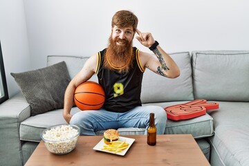 Caucasian man with long beard holding basketball ball cheering tv game smiling pointing to head...