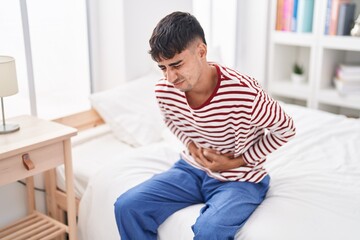 Young hispanic man suffering for stomach ache sitting on bed at bedroom