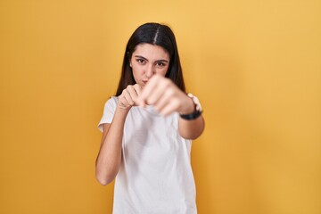 Young beautiful woman standing over yellow background punching fist to fight, aggressive and angry...