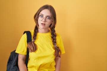 Young caucasian woman wearing student backpack over yellow background in shock face, looking skeptical and sarcastic, surprised with open mouth